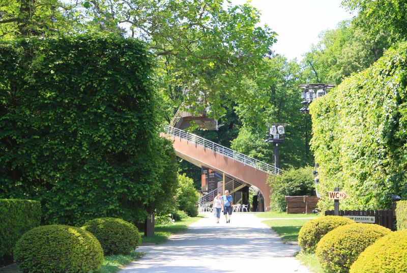 Two people walking along a path in leady Cesky Krumlov Gardens, with a revolving theatre in the background, including a cafe. 